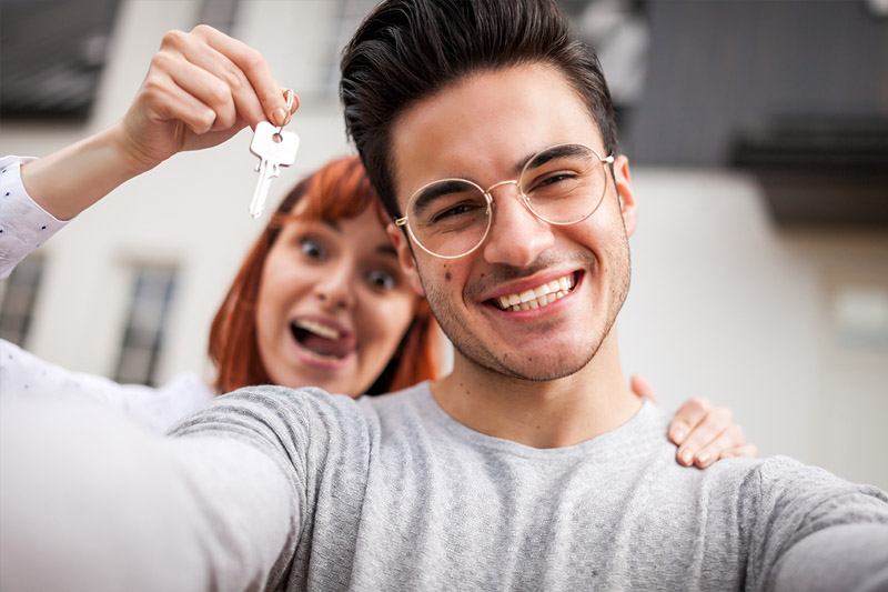 First Time Home Buyer 1 A smiling young man takes a selfie while a woman behind him excitedly holds up keys, suggesting they have just acquired a new home. Both appear happy and celebratory.