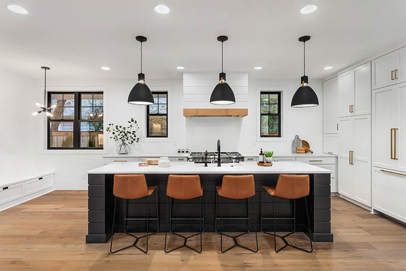 Jumbo Home Loan 1 Modern kitchen with a large white island, four brown barstools, black pendant lights, white cabinets, wood floor, and black-framed windows letting in natural light.