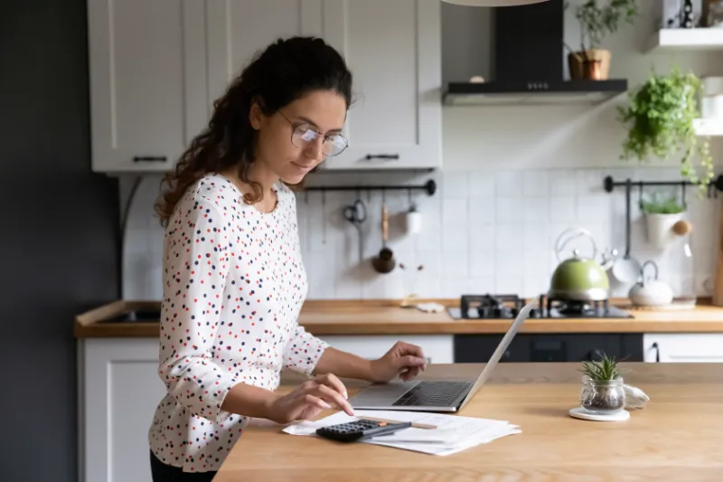 A woman stands in a kitchen, using a laptop and a calculator, with papers spread out on the counter. She appears focused, wearing glasses and a polka-dot shirt. Plants and kitchenware are visible in the background.
