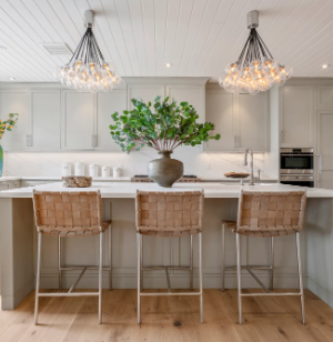 Modern kitchen with a large island, three woven bar stools, a vase of green branches as a centerpiece, pendant lights above, and light gray cabinets in the background.